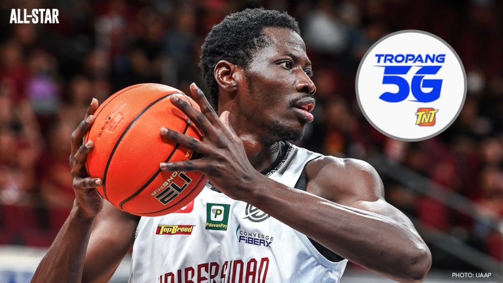 Malick Diouf holds a basketball while wearing a UP Fighting Maroons jersey as TNT Tropang 5G branding appears beside him ahead of the PBA semifinals.
